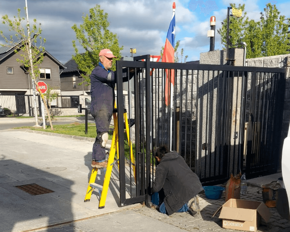 Dos técnicos de PROTEF instalando un portón automático: uno en la escalera y otro arrodillado en el acceso
