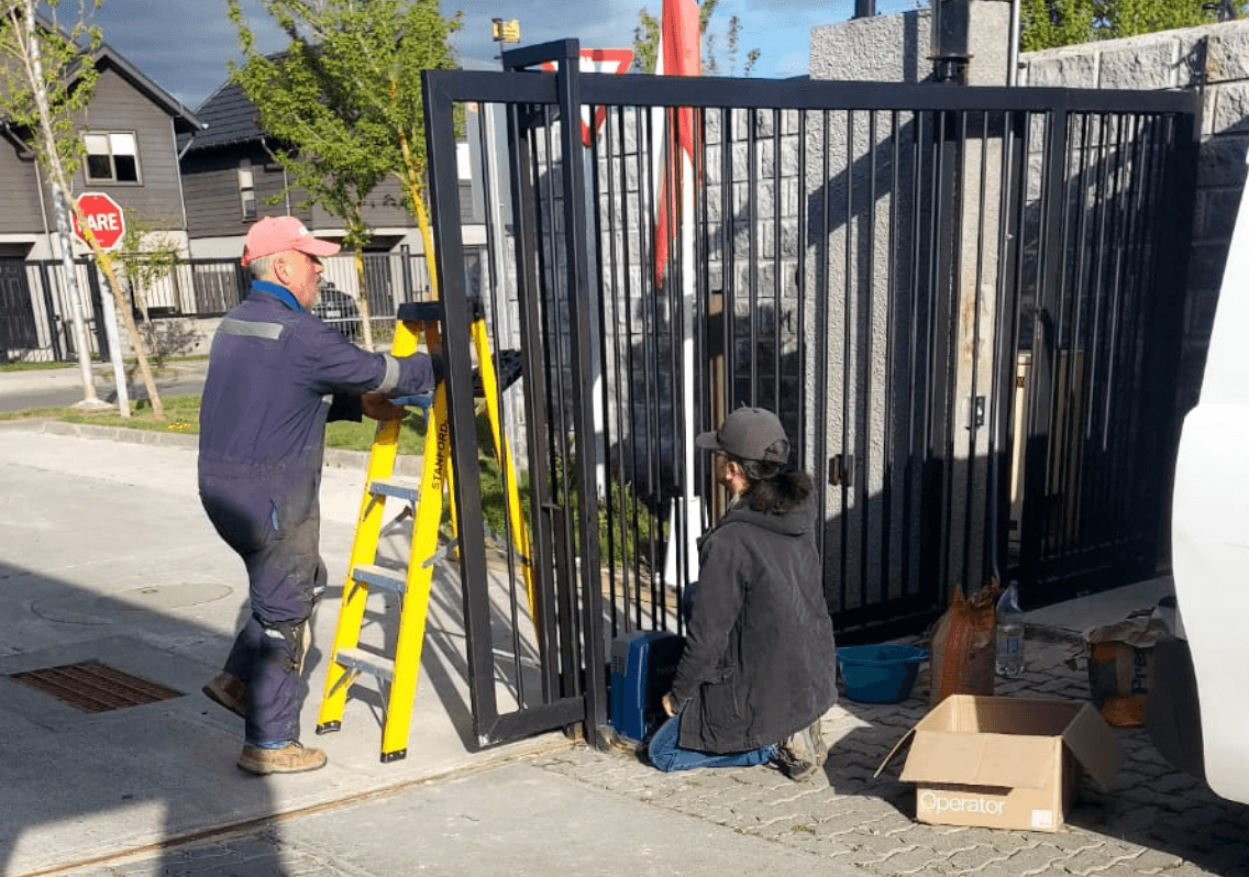 Dos técnicos de PROTEF instalando un portón: uno en la escalera y otro arrodillado junto al motor